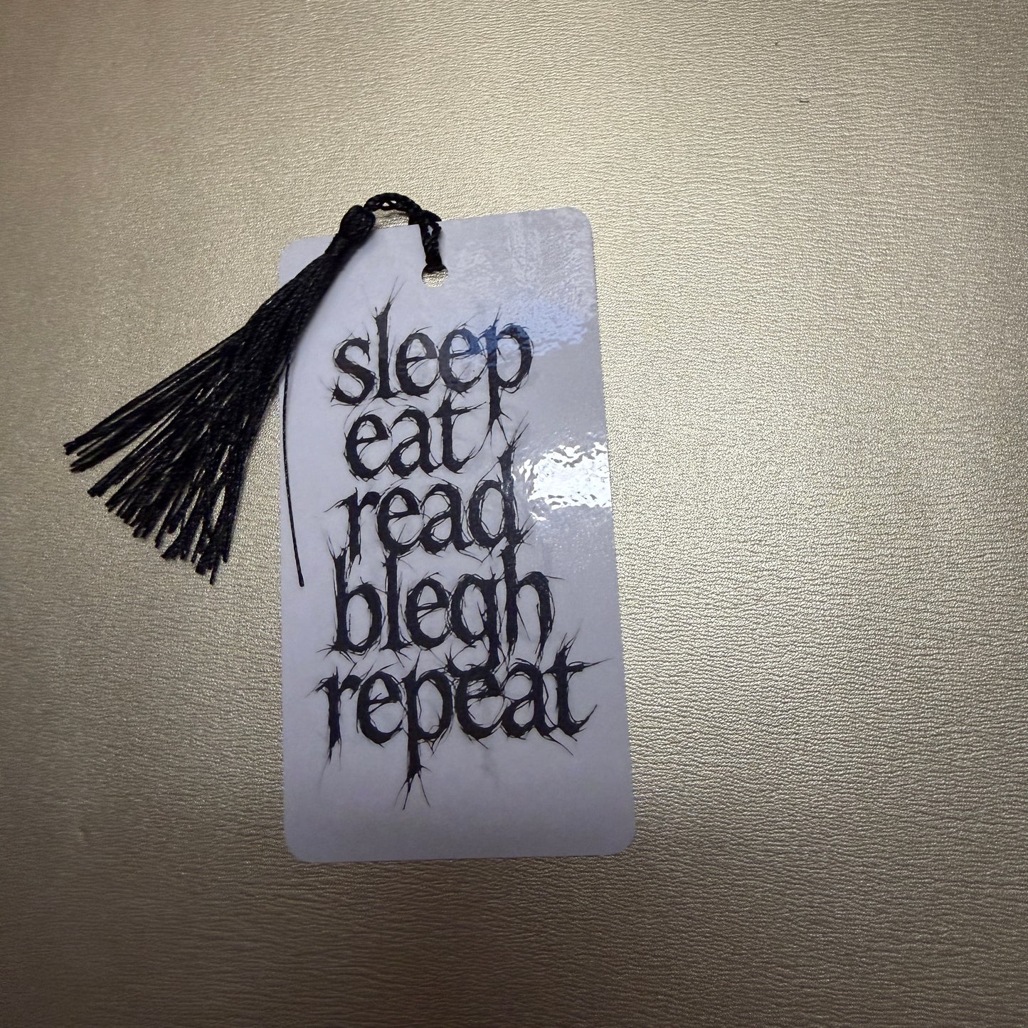 Weißes Lesezeichen mit schwarzen Quaste mit dem Schriftzug "sleep, eat, read, blech, repeat".