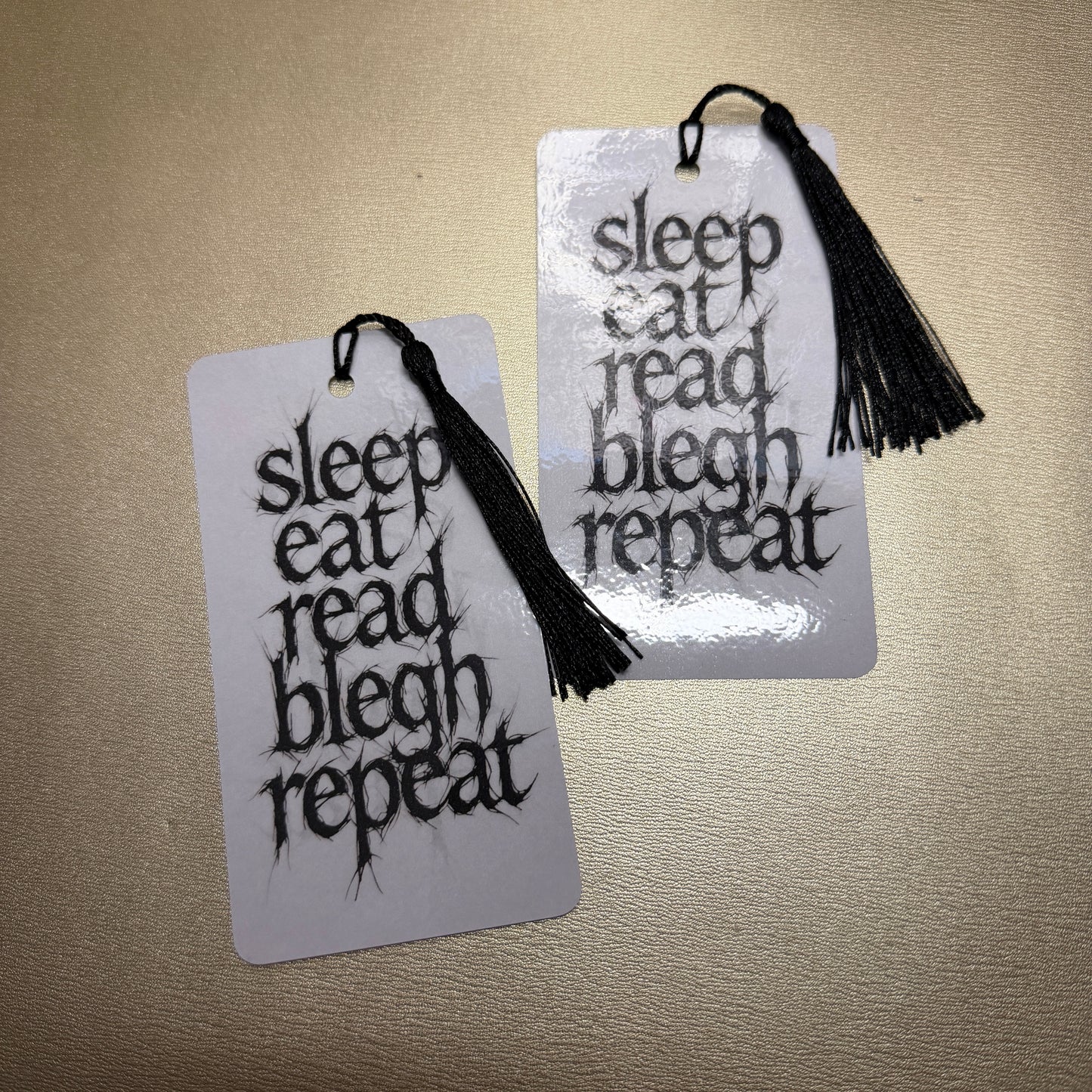 Weiße Lesezeichen mit schwarzen Quaste mit dem Schriftzug "sleep, eat, read, blech, repeat", Zwei Stück auf dem Bild.